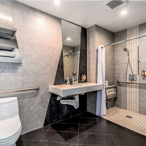 A modern bathroom with a toilet, mirror, sink, shower with grab bars, towel racks, and bottles of toiletries on the wall, all in a gray tile setting.