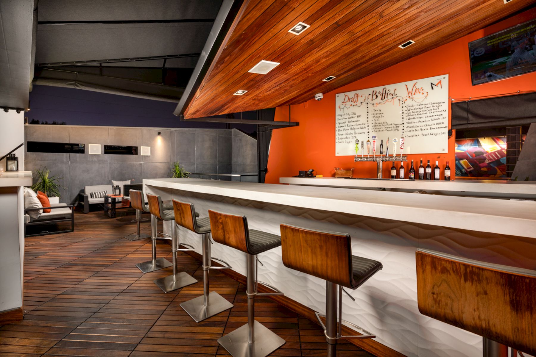 This image shows a sleek, modern bar with wooden barstools, a white countertop, and a menu on the orange wall. The seating area is visible in the background.