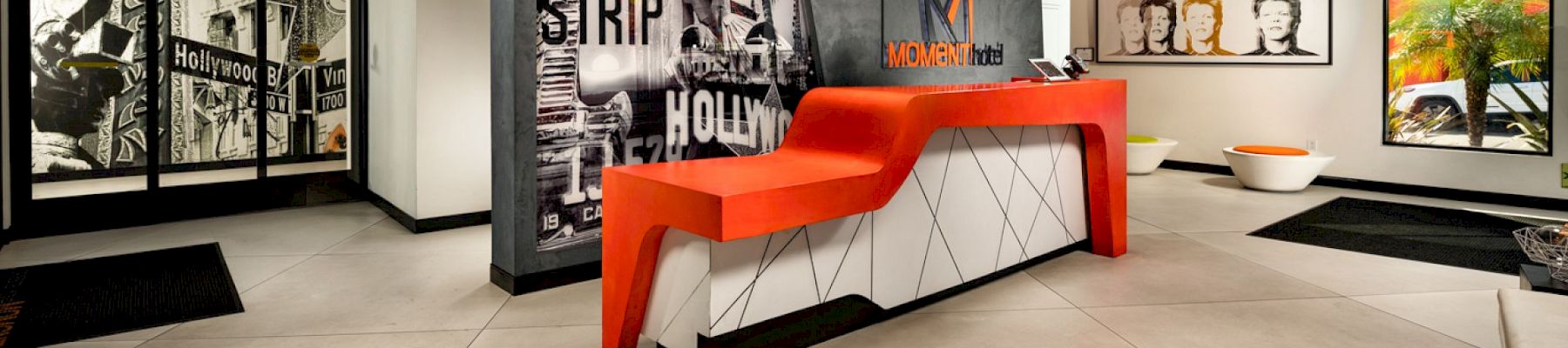 The image shows a modern hotel lobby with a red and white reception desk, art on the walls, and stylish lighting.