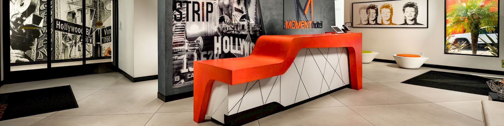 The image shows a modern hotel lobby with a red and white reception desk, art on the walls, and stylish lighting.