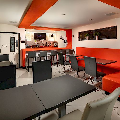 Interior of a modern dining area with black tables, red benches, and a bar in the background.